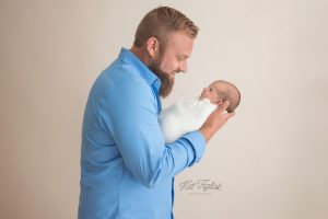 new dad holding his newborn baby girl wearing a blue shirt with a tan background