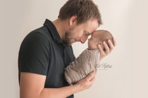 dad wearing black shirt holding newborn baby boy wrapped in tan knit wrap