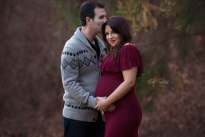expecting couple holding hands dad to be kissing his wife pregnant mom is wearing a cranberry off the shoulder dress