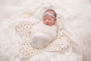 Newborn baby girl smiling and swaddled in white wrap with floral headband on white rug