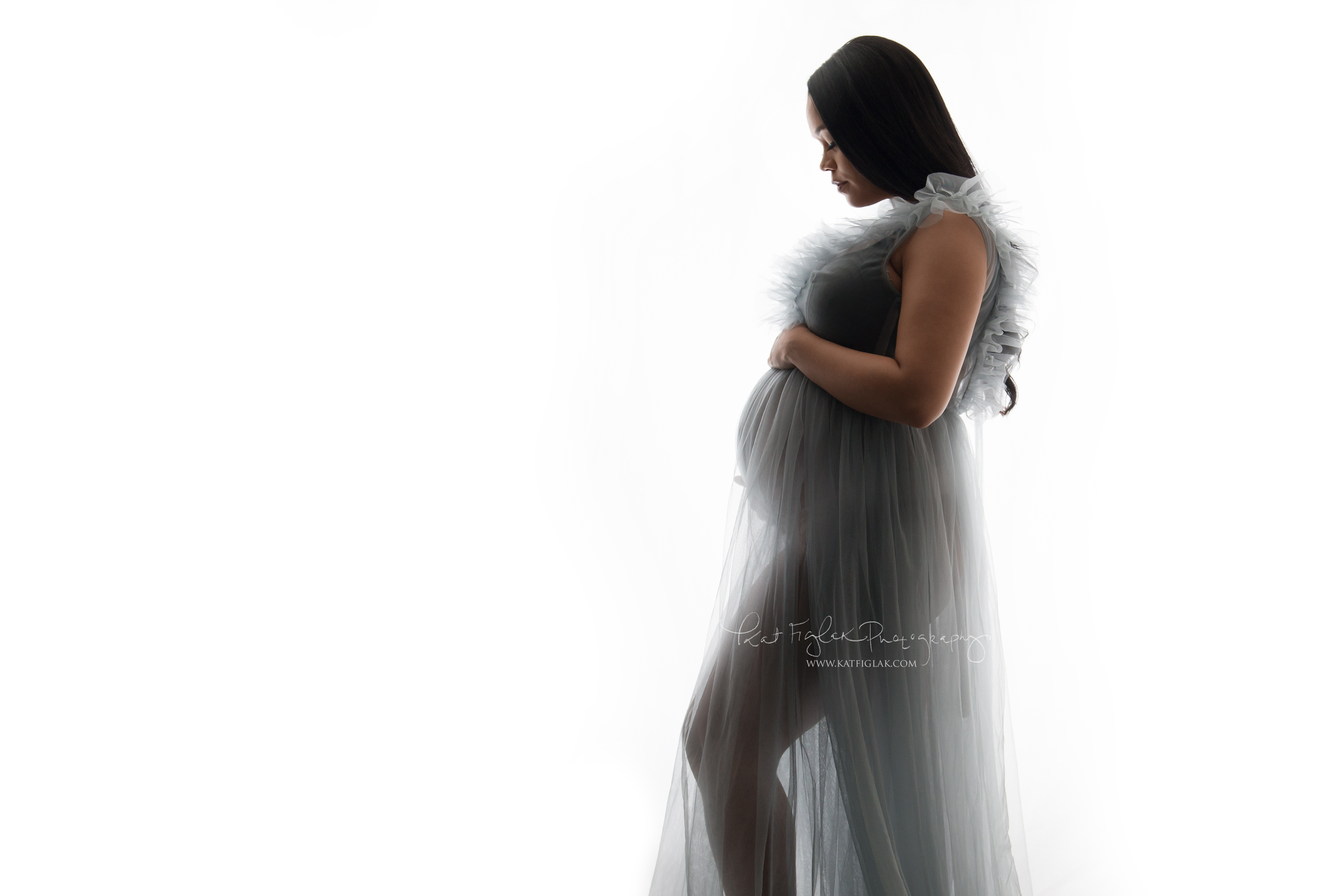 Pregnant woman standing looking at her belly wearing a shear gray gown with light coming in from the background