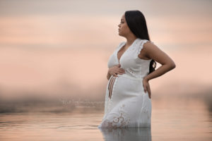 Pregnant woman standing in water with a warm sunset behind her wearing a white dress