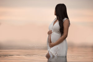 Pregnant woman standing in lake holding her belly looking off at the horizon sunset