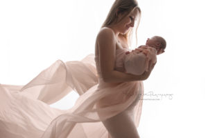 Mom holding her newborn baby girl looking at her wearing a blush gown that is flowing behind her