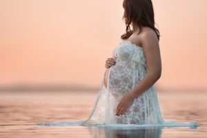 Pregnant woman standing in the lake looking out at the horizon wearing a white lace dress