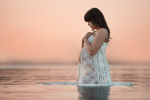 pregnant woman standing in lake wearing a white lace dress looking at her belly with a beautiful sunset in the background