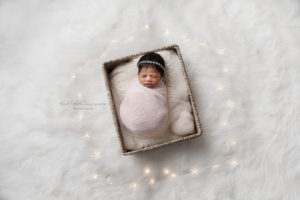 Newborn baby girl wrapped in blush wrap sleeping in a basket on white fur with star holiday lights around the basket
