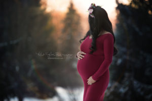 pregnant mom looking off in the distance of evergreens wearing a ruby red dress and floral crown