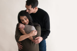 Mom holding her newborn son wearing olive green being kissed by her husband wearing a black sweater