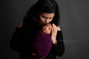 mom holding her newborn baby girl and kissing her on the forehead baby is wrapped in a purple wrap