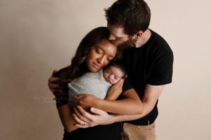 mom holding baby boy wrapped in gray wrap with dads arms around them kissing mom