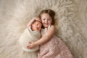 Little girl in a fancy pink dress holding her newborn baby sister who is wrapped in a cream knit wrap.