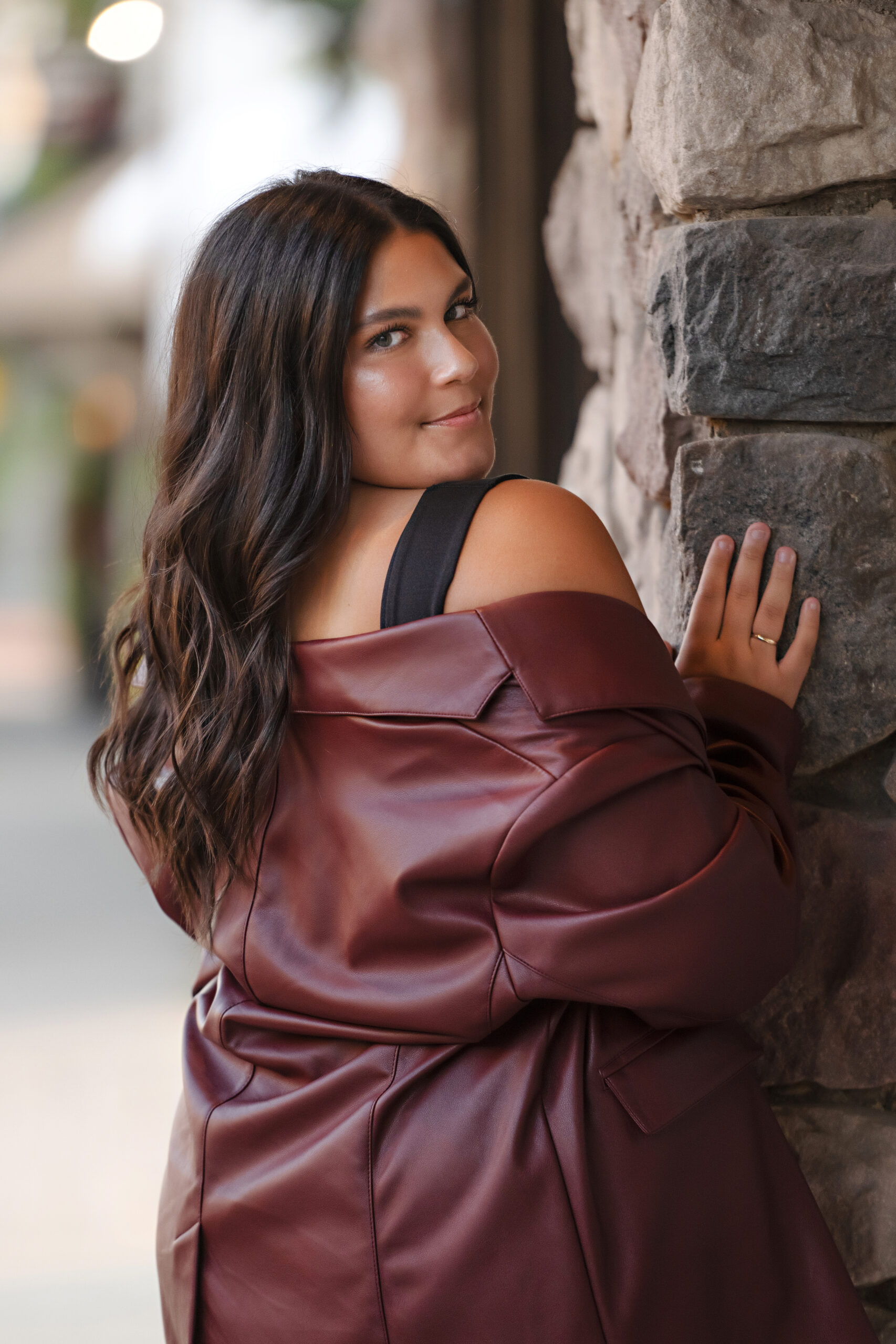 High school senior girl who has dark brown hair facing a stone wall waring a black tank top with a maroon leather jacket draped off her shoulders