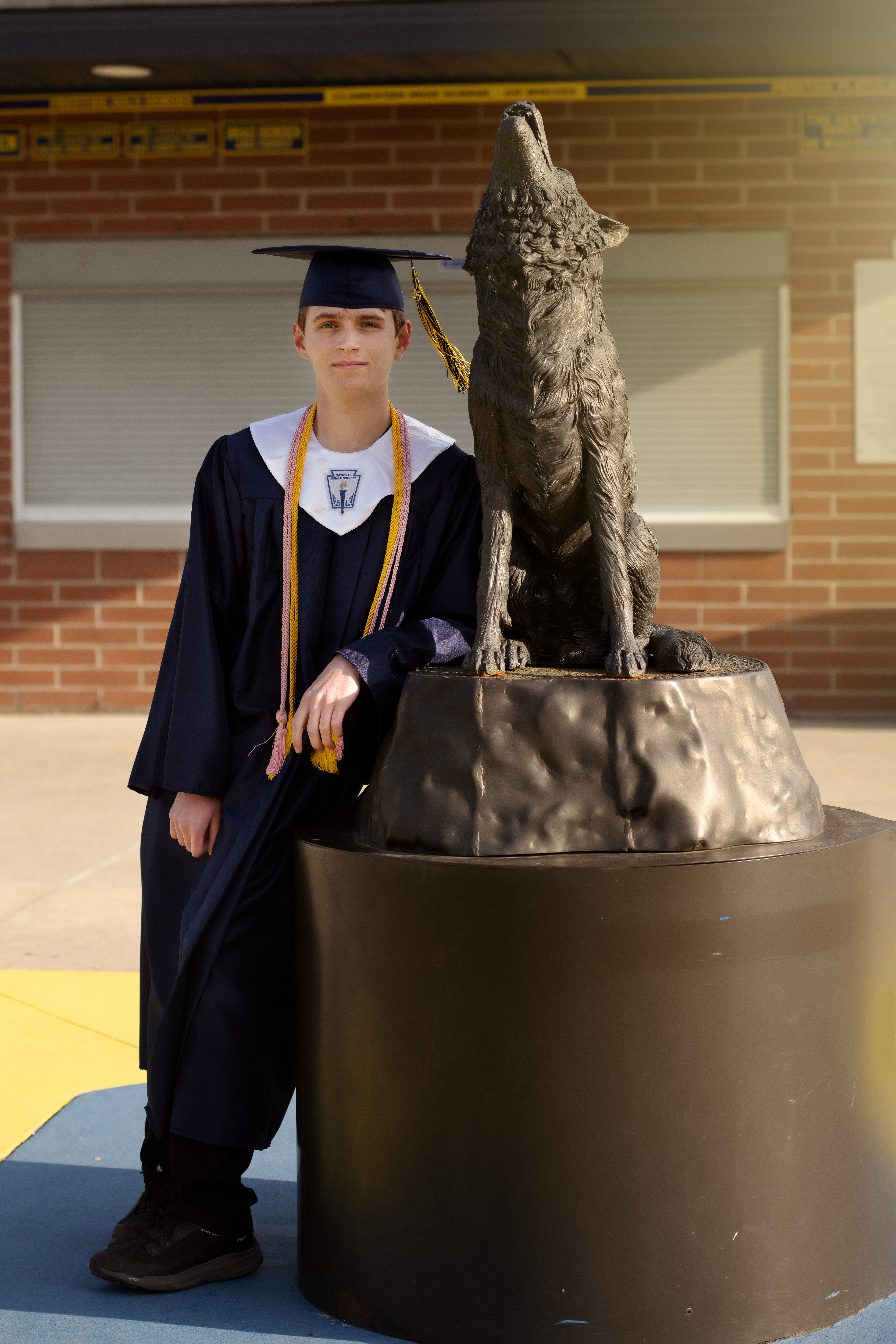 senior boy wearing his navy blue cap and gown for graduation standing next to the Clarkston high school wolf in the football stadium