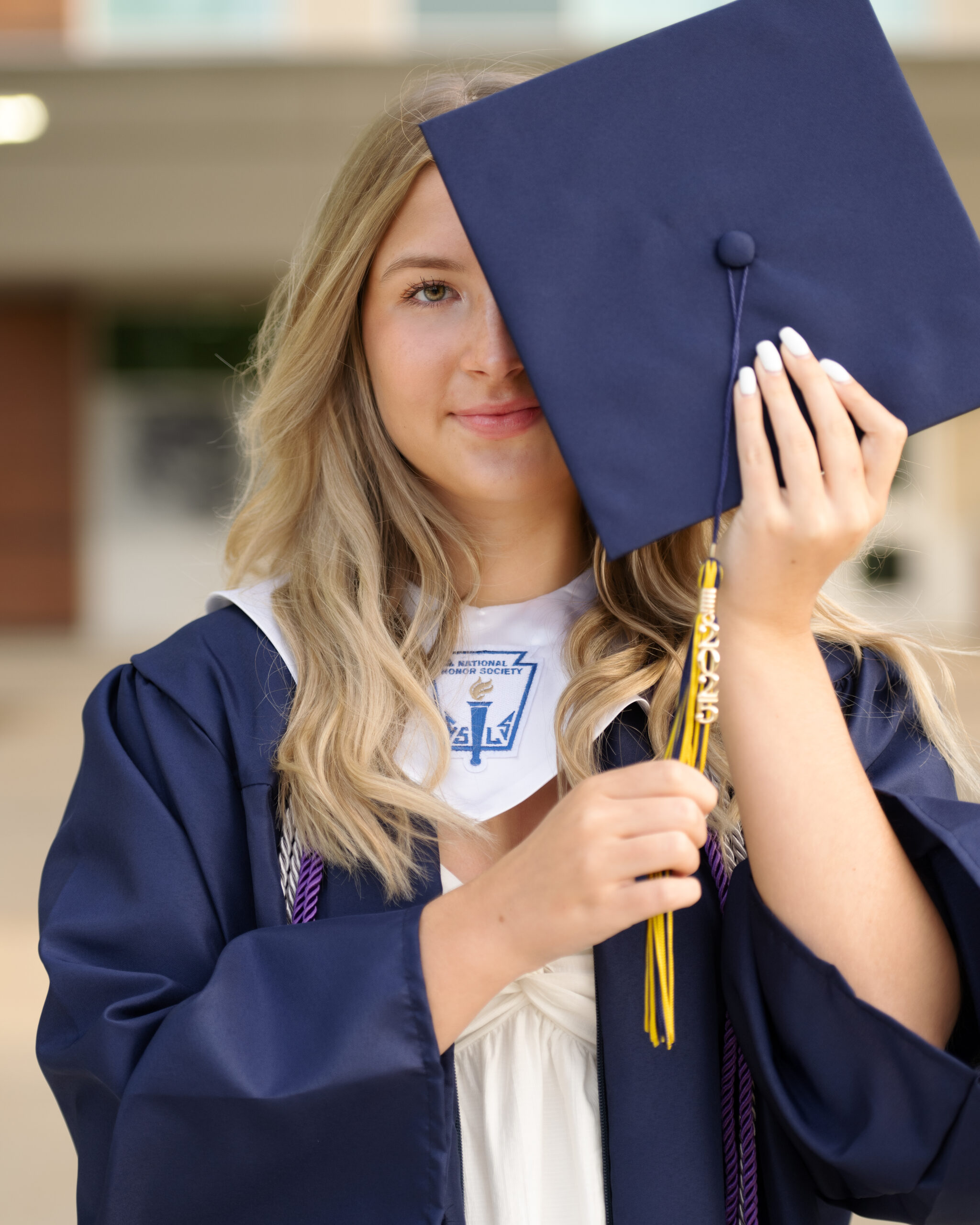 female clarkston high school grad wearing navy graduation cap and gown  holding her cap half across her face with her long blonde hair pulled forward