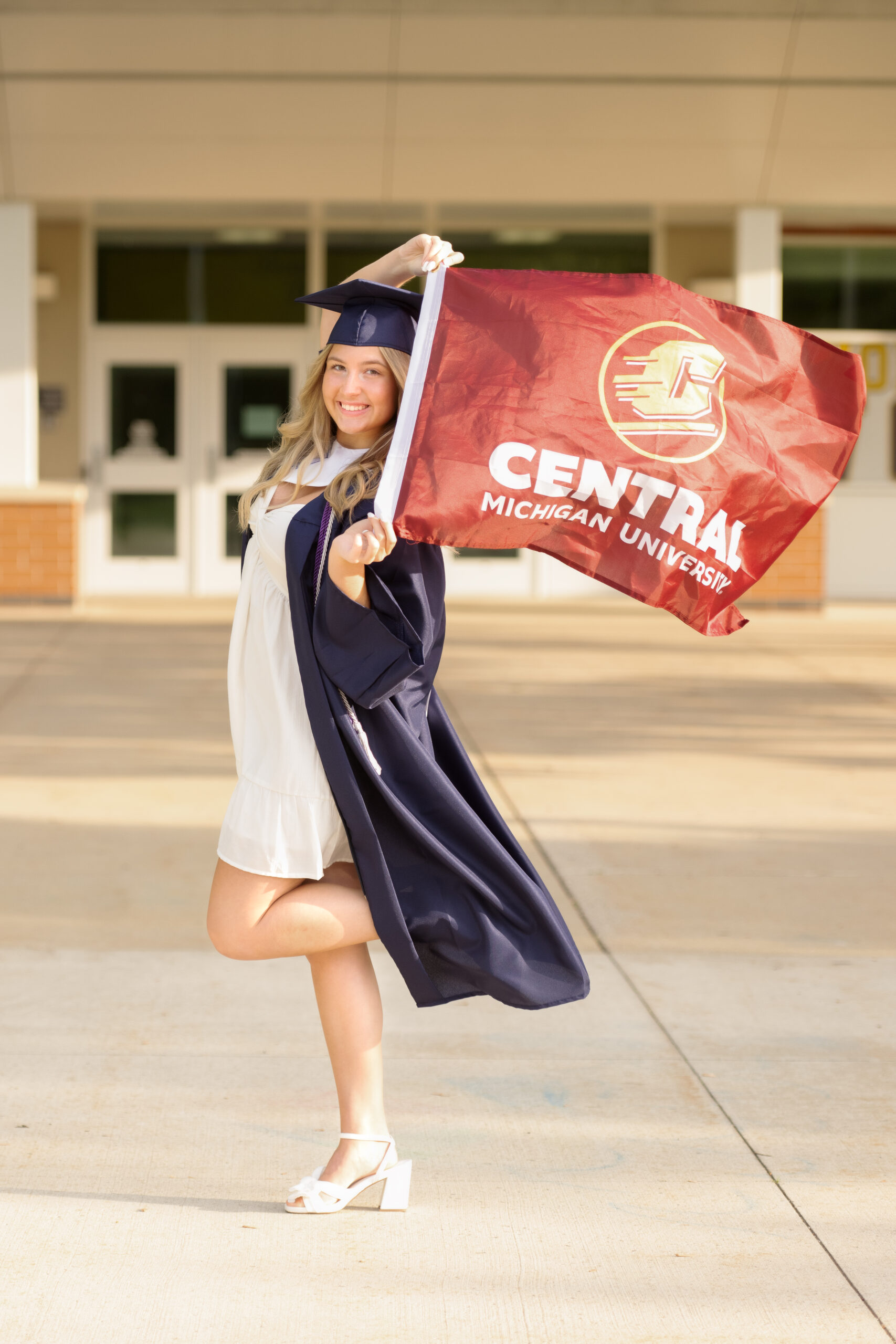 female clarkston high school grad wearing navy graduation cap and gown  with a white dress under holding a central university flag above her head