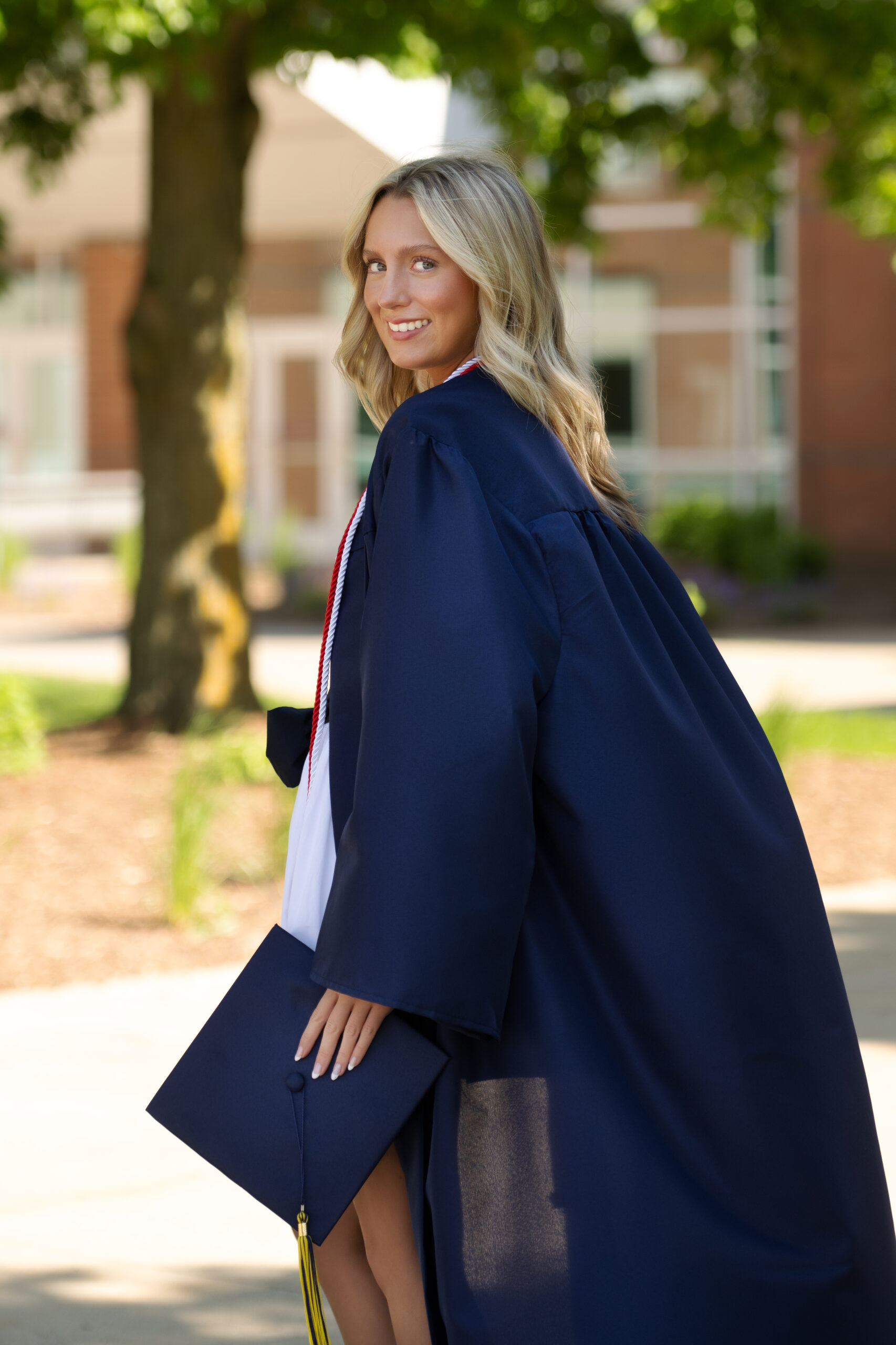 female clarkston high school grad wearing navy graduation cap and gown  standing with her gown blowing behind her in front of Clarkston High school