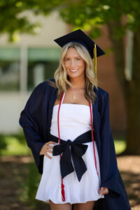 female clarkston high school grad wearing navy graduation cap and gown standing with her hands on her hips standing in front of clarkston high school