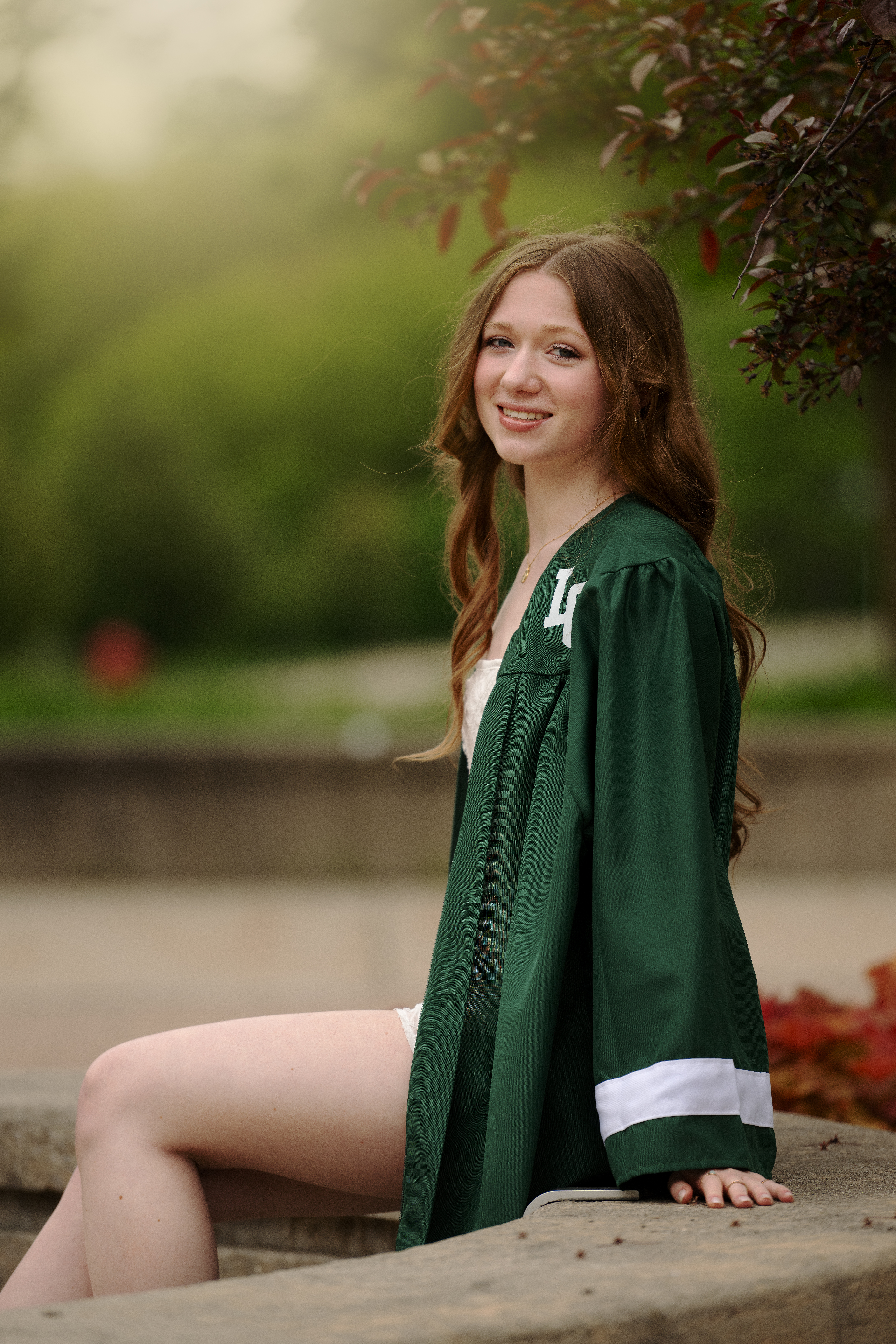 Lake Orion female grad with long strawberry blonde hair wearing her forest green cap and gown sitting on a stone bench in front of Lake Orion High school