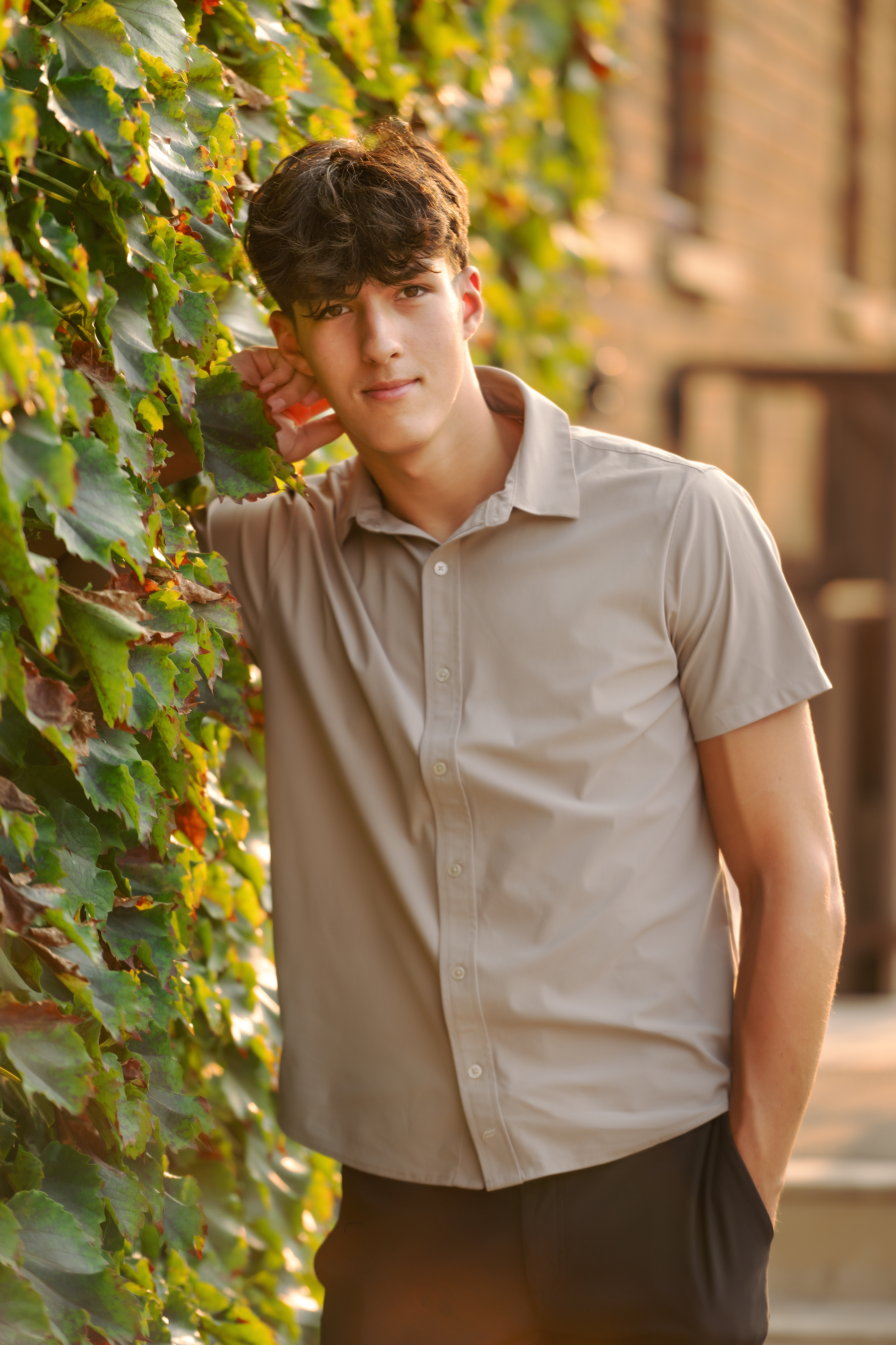 Flint Powers Catholic Senior boy wearing a tan shirt with dark hair leaning against an ivy wall in downtown Clarkston, MI