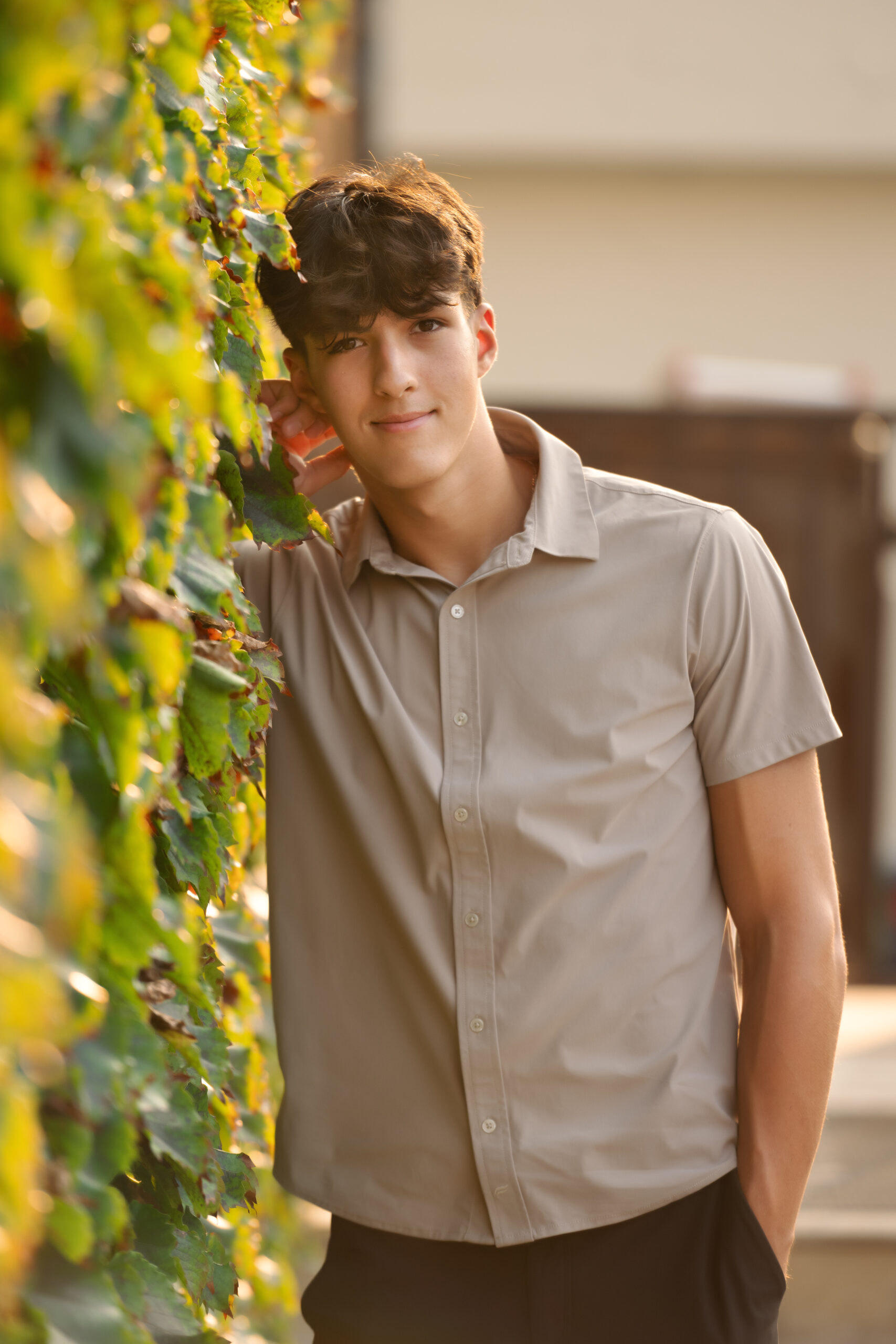 young senior boy wearing a tan shirt and black pants leaning up against a wall of green ivy in downtown Clarkston
