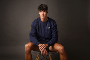 Flint Powers Catholic Senior boy sitting on a stool wearing a navy Nike sweatshirt with a dark backdrop