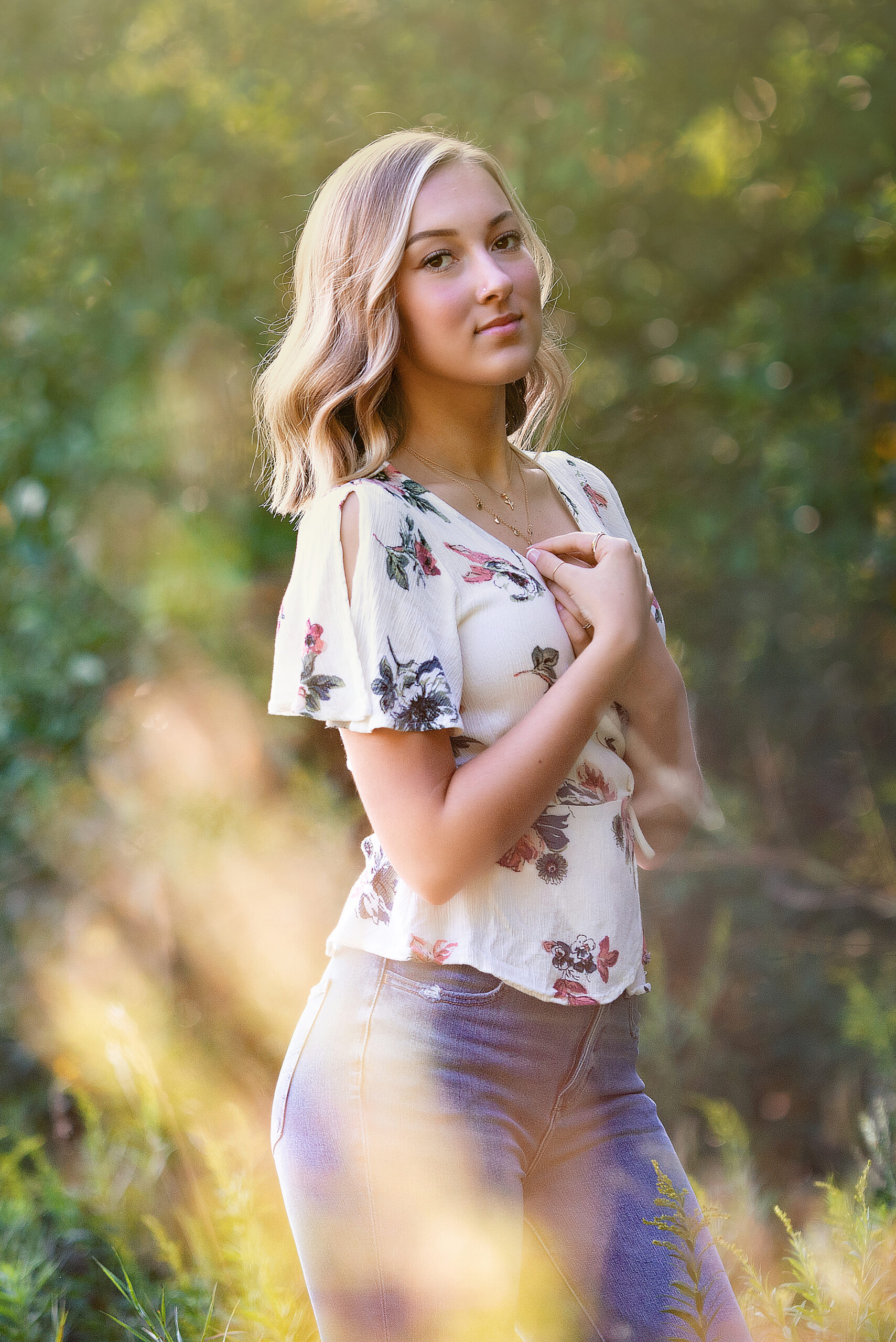 Girl standing in a field with the sunlight kissing the back of her head she has blonde shoulder length hair and is wearing a floral short sleeve shirt and jeans