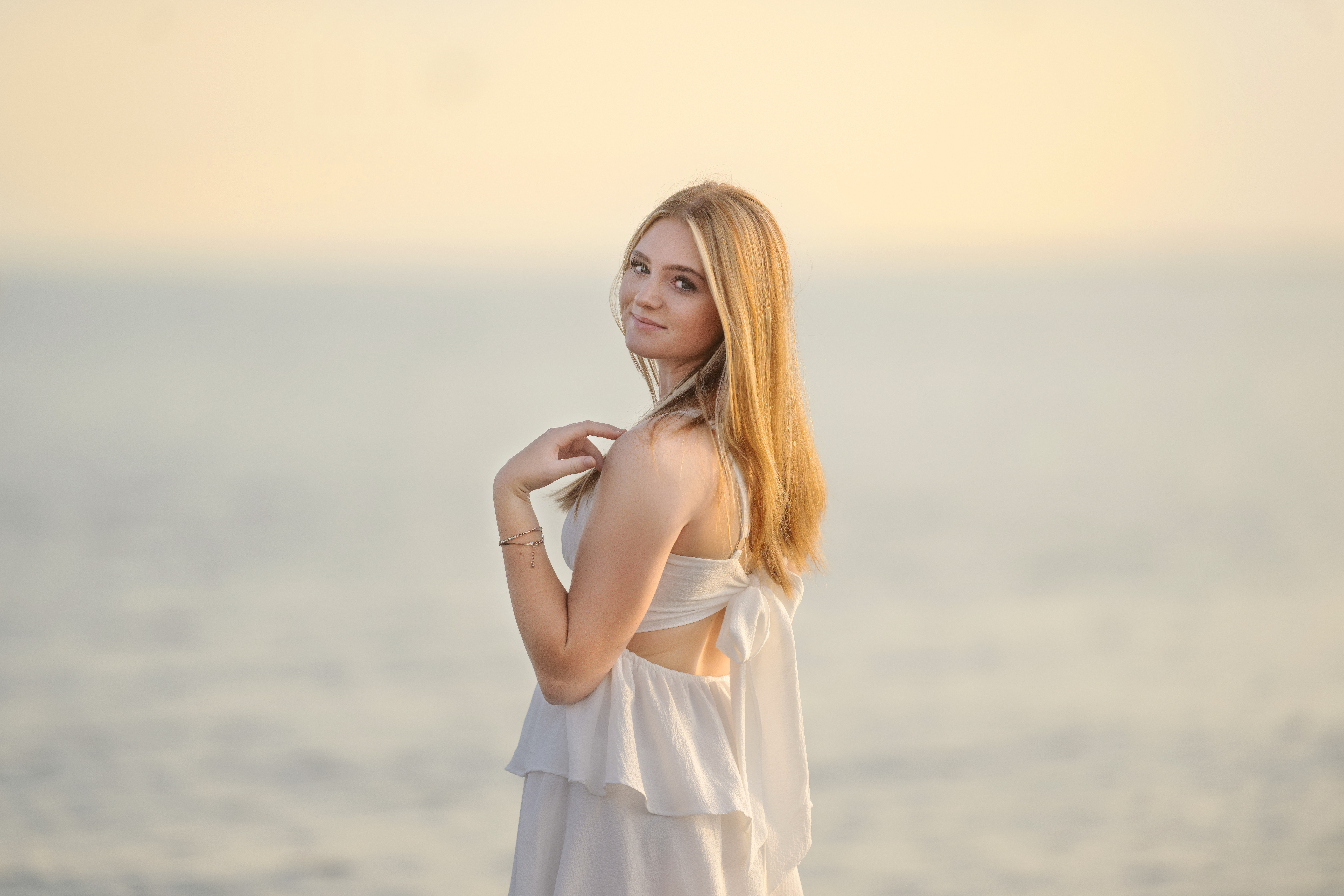 young senior girl with auburn hair with blonde highlights standing in the lake wearing a white ruffled dress.