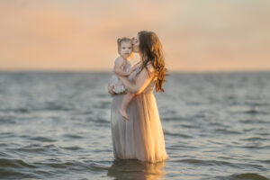 photo of a pregnant woman wearing a tan tulle gown standing in Lake St. Clair holding her toddler daughter who is wearing a pink tutu