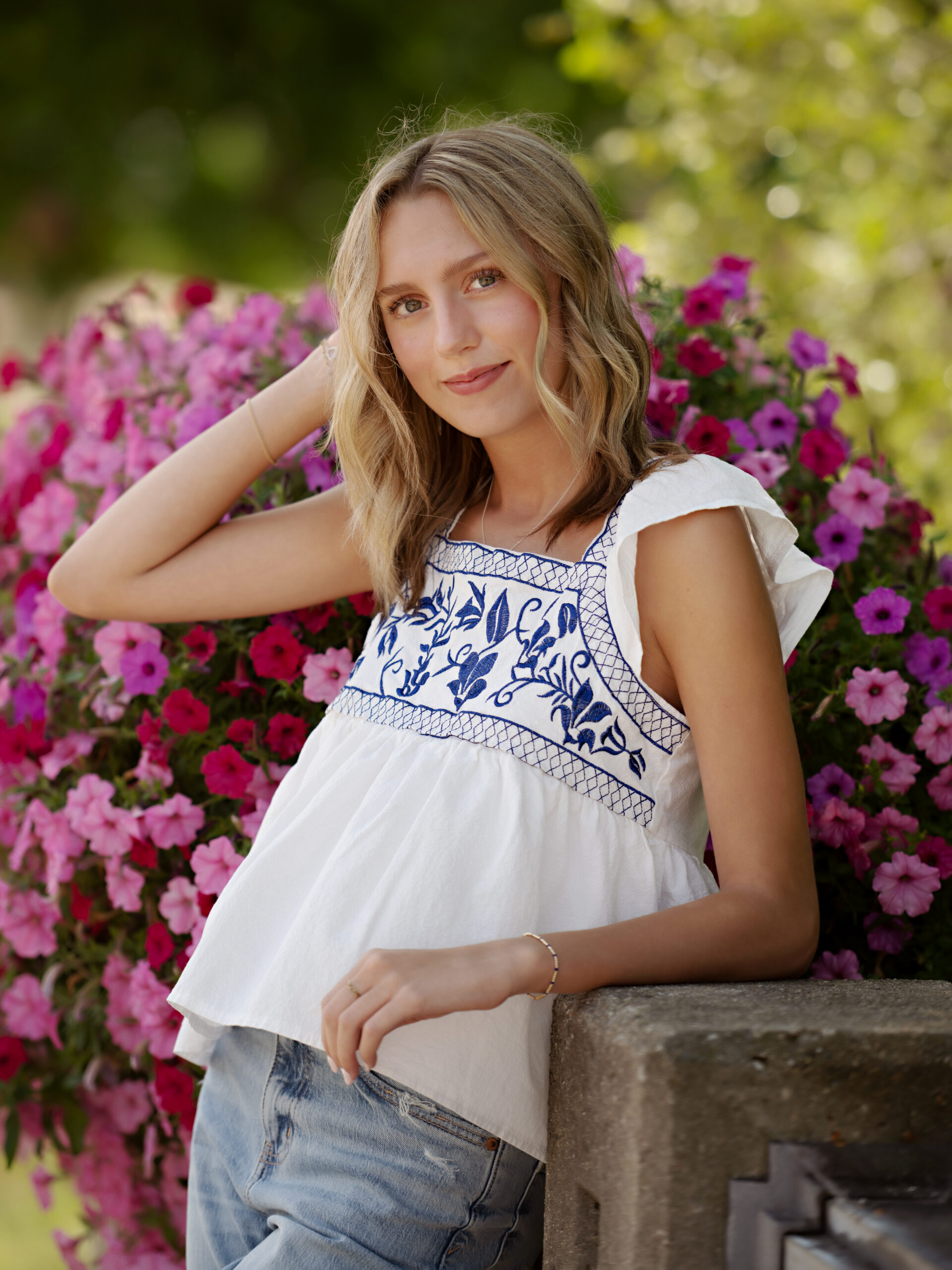 Photo of a high school senior girl wearing a white top with blue floral embroidery. Standing in front of purple flowers