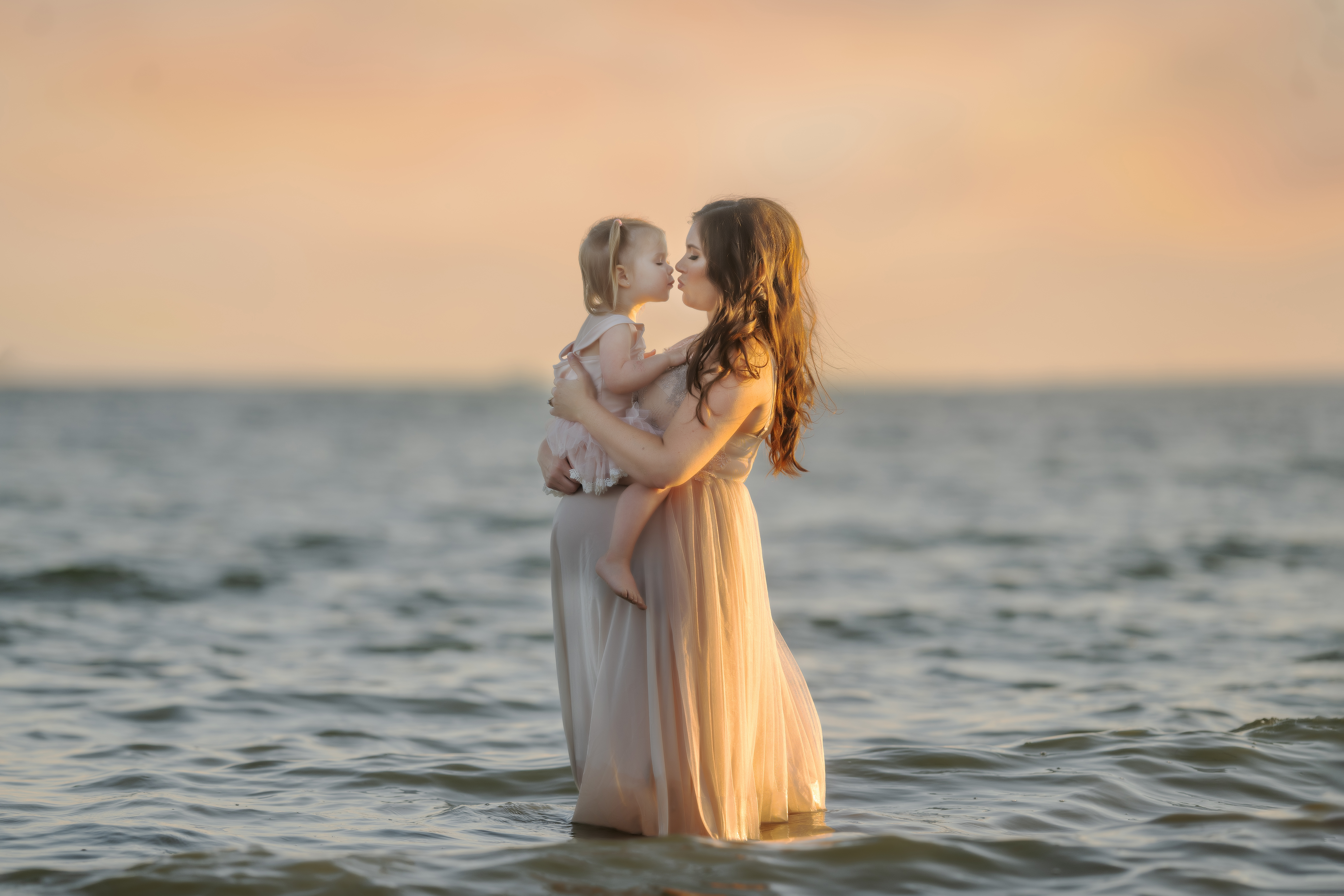 photo of a pregnant woman wearing a tan tulle gown standing in Lake St. Clair holding her toddler daughter who is wearing a pink tutu
